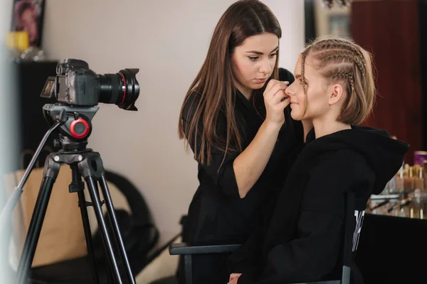 Side view of make up artist work with beautiful blond hair model in studio until camera films process of makeup. Blog of makeup artist