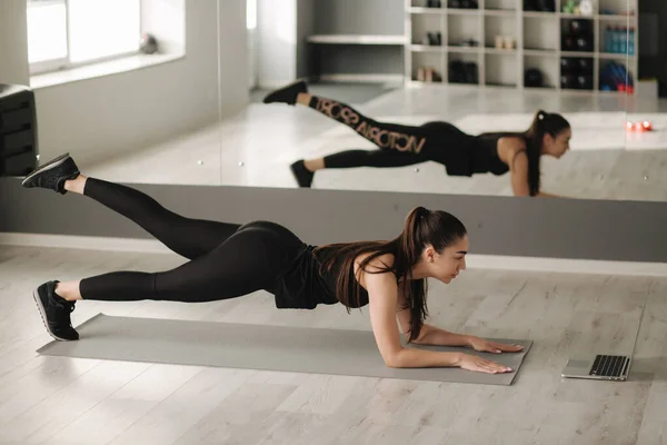 Atractiva mujer en forma haciendo tablón de yoga mientras ve tutoriales en línea en el portátil. Formación solo en estudios —  Fotos de Stock