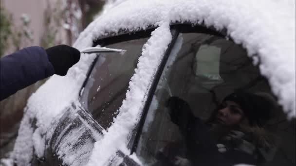 Une femme nettoie une vitre enneigée sur une voiture avec un grattoir à neige. Jolie femme chaude habillée nettoyer sa voiture à l'extérieur. Froid matinée enneigée et givrée. Voiture noire — Video