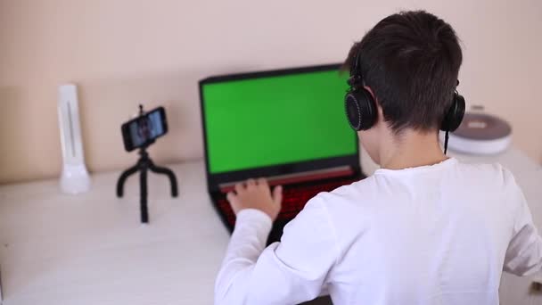 Vista posteriore di giovane ragazzo gioco sul computer portatile. Schermo verde nel computer portatile. Steamer in auricolare cattura video sul suo telefono su treppiede. Scherzi? — Video Stock