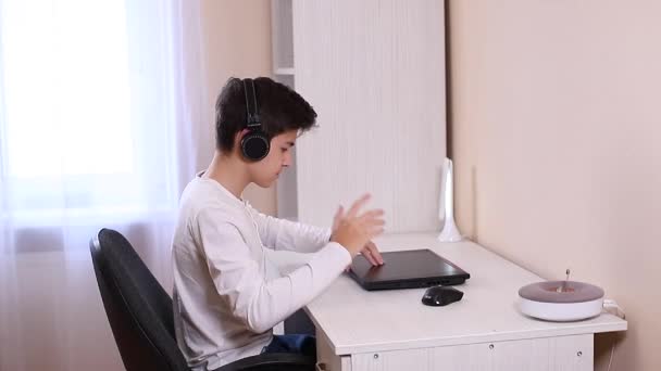 Мальчик в наушниках открывает ноутбук и начинает играть в игры. Красавчик-подросток — стоковое видео