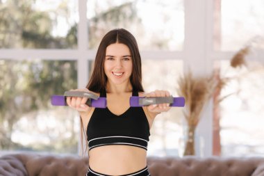 Evde genç ve çekici bir bayan antrenmanı. Kız bikini sezonuna hazırlanıyor. Mutlu kadın eğitim için dambıl kullanır.