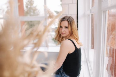 Stüdyoda genç ve çekici bir kadın pencerenin yanında oturuyor. Kot giymiş bir kadın. Ev biçimi