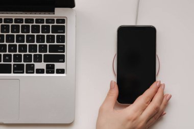 Female hand tup phone on wireless charging. Work place near at the laptop. Top view. Place for text