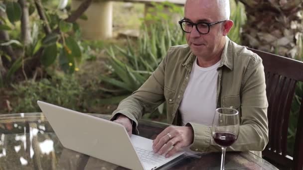 Middle age man sitting outdoors by the table and working on laptop. Man drink dry red wine during the work on backyard. Freelancer on quarantine. — Video