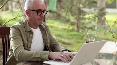 Middle age man sitting outdoors by the table and working on laptop. Man drink dry red wine during the work on backyard. Freelancer on quarantine.