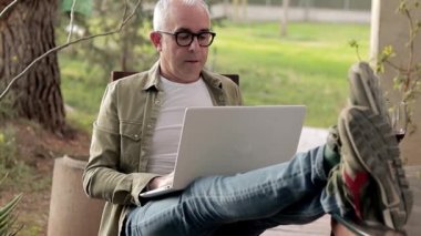 Gray-haired middle aged man using laptop outdoors. Man put feet on table and relax during a break