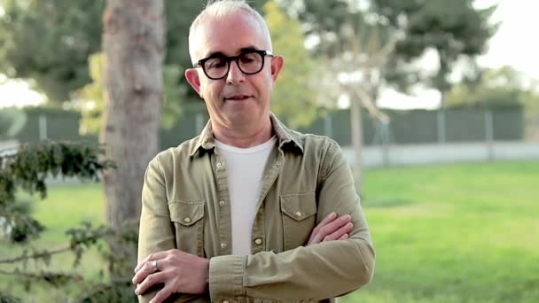 Gray-haired middle aged man in black glasses stand in front of green background and look in the camera. Man standing outdoors and smile — Wideo stockowe