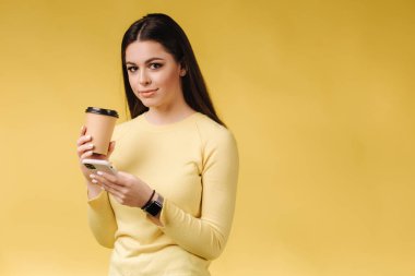 Pretty girl using phone and drink coffee on yellow background. Studio photoshoot of brunette female in yellow sweater. Happy smiled woman. Place for text