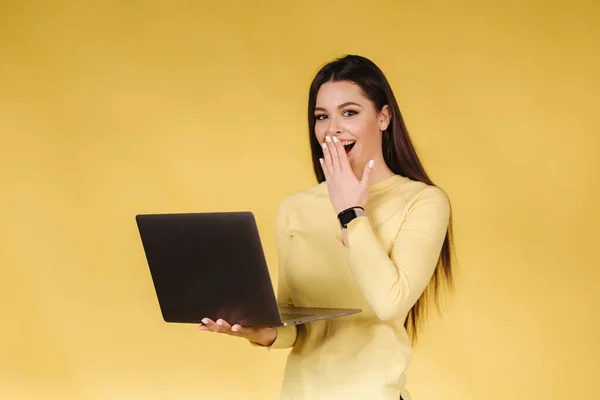 Happy surprised woman in yellow sweatshit hold laptop and raised hand to face. Portrait of pretty woman on yellow background. Pantone