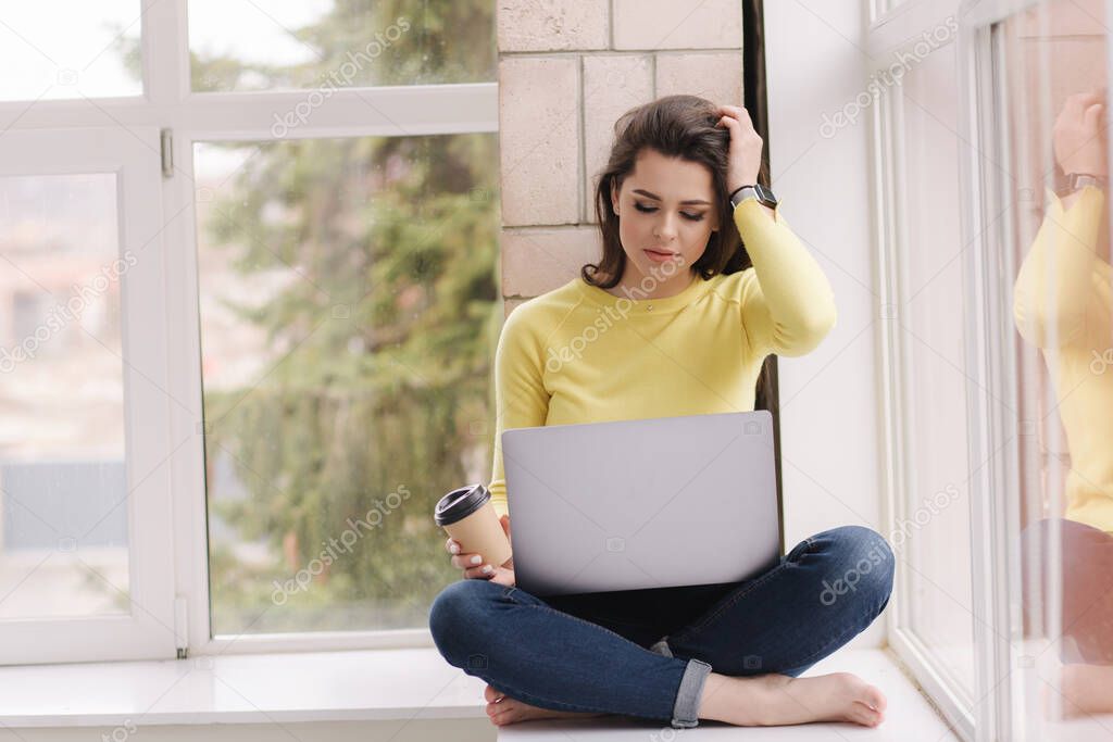 Young graphic designer working at home. Woman sitting on window sill with laptop and drink coffee. Self isolated female freelancer. Barefoot