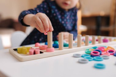 Çocukların eline ahşap kolona renk halkaları takması. Küçük kız masada çocuklar için eğitici bir oyun oynuyor. Farklı renkler ve sayılar içeren tahta bir oyun. Akıllı için intekaktif oyun