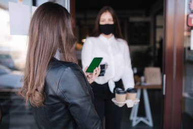 Kadın dışarıda kafenin yanında duruyor ve Covid-19 salgını sırasında akıllı telefonla ödeme yapıyor. Müşteri NFC ödemesi için telefon tutarken, kredi kartı okuyucu makinesini elinde tutan kasiyer. Telefondaki yeşil ekran