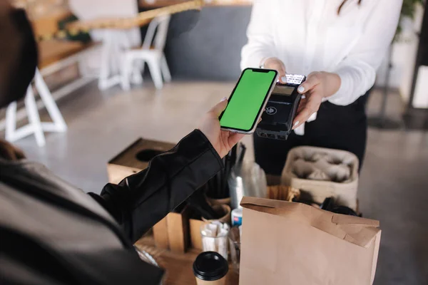 First person view of female paying with smartphone during Covid-19 pandemic. Cashier hand holding credit card reader machine while client holding phone for NFC payment. Green screen on phone. Mock up