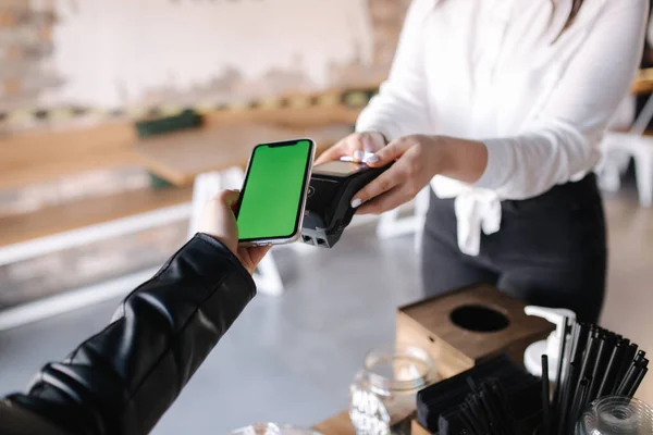 First person view of female paying with smartphone during Covid-19 pandemic. Cashier hand holding credit card reader machine while client holding phone for NFC payment. Green screen on phone. Mock up