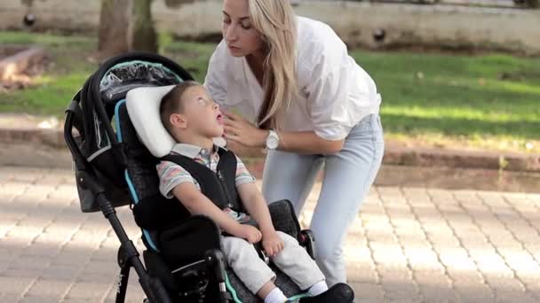 Видео счастливой семейной матери, гуляющей на улице и помогающей смотреть на сына-инвалида, сидящего в инвалидной коляске. Концепция ухода за инвалидами — стоковое видео