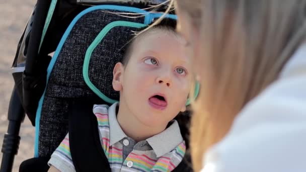把小孩关在外面的轮椅上身体残疾的孩子和妈妈一起在公园里散步。照顾她儿子的单亲父母 — 图库视频影像
