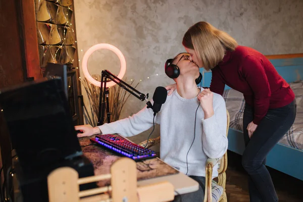 Young attractive wife help husband to work. Handsome man sits at the table and work on computer. Man using professional microphone and headphones. Woman kiss man