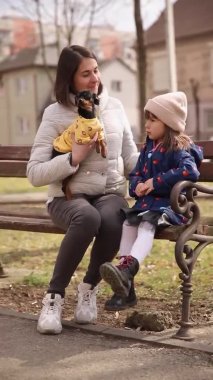 Annemin kızı ve küçük köpeğiyle bankta oturduğu dikey video. Mutlu aile dışarıda yürüyor.
