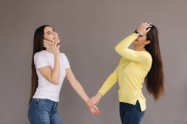 Bir kız telefonla konuşur, diğeri şaşırır ve kızar. Stüdyoda gri arka planda çekici genç kadınlar var.