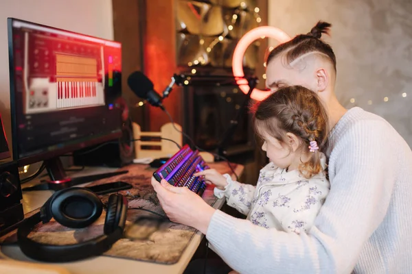 Younger sister sits on brothers knees and put finger on keyboard with neon light. Little girl help brother work on computer. Happy family