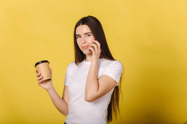 Portretul unei fete atractive vorbind la telefon și țineți o ceașcă de cafea pe fundal galben. Locul pentru text. Privind camera . — Fotografie, imagine de stoc