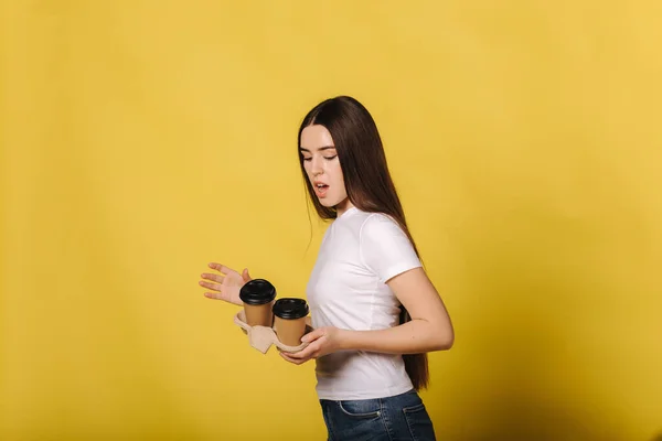 Female eccidentaly fall down two cups of coffee. Woman arfaid of coffee drops on the floor. Woman in white t-shirt standing isolated over yellow background