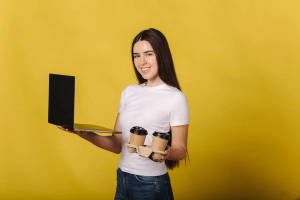 Retrato de mujer morena usando laptop y sostener dos taza de capuchino sobre fondo amarillo en el estudio. Mujer con camiseta blanca trabajando y pensando. Mujer feliz sonrisa y mira al fotógrafo. Lugar para —  Fotos de Stock