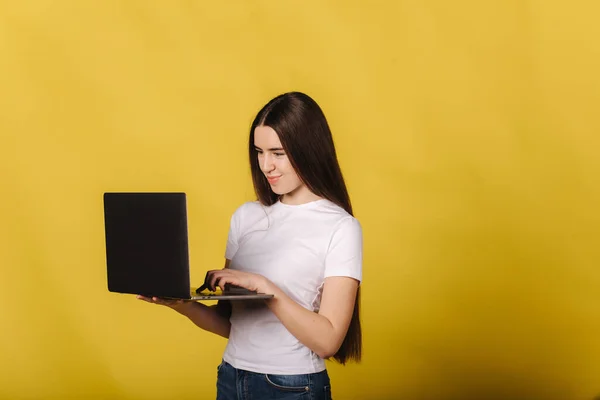 Πορτρέτο της μελαχρινής γυναίκας χρησιμοποιώντας φορητό υπολογιστή σε κίτρινο φόντο στο στούντιο. Γυναίκα με λευκό μπλουζάκι που δουλεύει και σκέφτεται. Ευτυχισμένη γυναίκα χαμογέλασε και ψάξε στο λάπτοπ. Θέση για κείμενο — Φωτογραφία Αρχείου