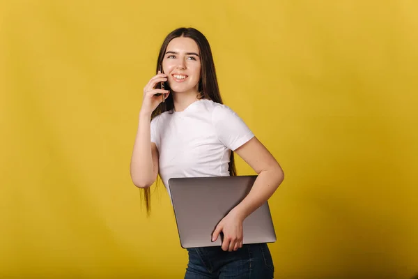 Ελκυστική γυναίκα με φανελάκι κρατάει κλειστό το λάπτοπ και μιλάει στο τηλέφωνο. Θέση για κείμενο. Γυναίκα στέκεται απομονωμένη πάνω από κίτρινο φόντο — Φωτογραφία Αρχείου