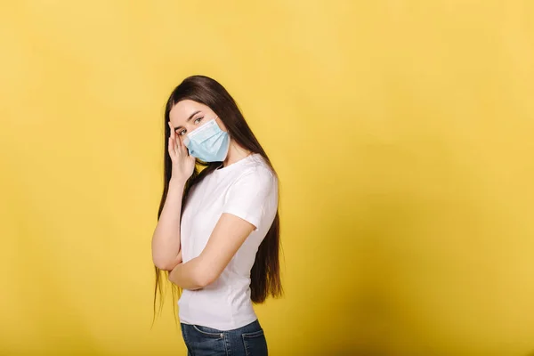 Tıbbi maskeli genç bir kadının portresi sarı arka planda duruyor. Kızın yüz maskesi yüzünden başı ağrıyor. Koruyucu maskeli bir kadın kafasını eliyle tutuyor. — Stok fotoğraf