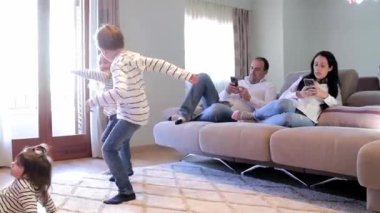 Anne ve baba kanepeye oturur ve çocukları içeride oynarken telefon kullanırlar. Çocuklar birlikte vakit geçirirken aile blogcuları çalışıyor. Evde iki kız ve bir erkek.