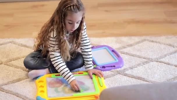 Mała dziewczynka bawi się w domu podczas kwarantanny. Dziewczyna siedzi na podłodze i maluje na tabliczce z zabawkami — Wideo stockowe