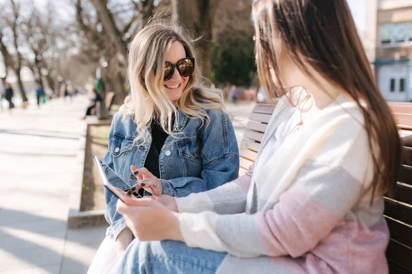 Twee vrouwelijke vrienden zitten op de bank in het park en zoeken iets in tabletvorm. Jonge en gelukkige meisjes in het voorjaar kleding online kopen — Stockfoto
