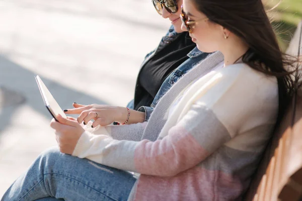 Twee vrouwelijke vrienden zitten op de bank in het park en zoeken iets in tabletvorm. Jonge en gelukkige meisjes in het voorjaar kleding online kopen — Stockfoto