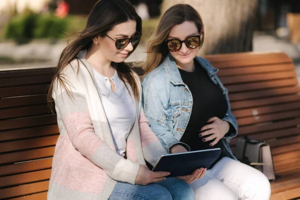 Schwangere mit ihrer besten Freundin sitzt im Park und sucht per Tablet nach einem Kinderwagen. Zwei Frauen mit Sonnenbrille wählen Kleidung für Baby — Stockfoto