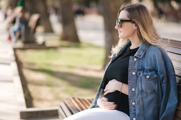 Feliz mujer embarazada poner las manos en el vientre y mirando en el río. Mujer sentada en el banco en el parque. Futuro aliento de madre aire fresco — Foto de Stock