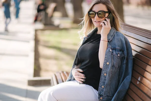 Mujer embarazada se sienta en el banco en el parque y el uso de teléfono. Feliz hembra descansar al aire libre durante los paseos — Foto de Stock