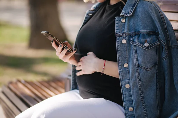 La selección media de la mujer embarazada se sienta en el banco en el parque y el uso de teléfono. Feliz hembra descansar al aire libre durante los paseos — Foto de Stock