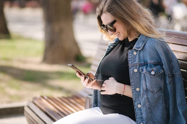 Pregnat vrouw zit op de bank in het park en het gebruik van telefoon. Gelukkig vrouwtje hebben rust buiten tijdens de wandelingen — Stockfoto