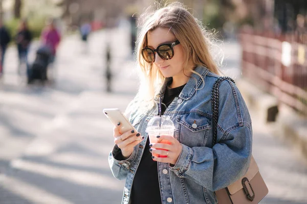Hübsche Frau, die Erdbeerlimonade trinkt, während sie draußen spaziert. Porträt einer attraktiven Frau mit Sonnenbrille am Telefon — Stockfoto
