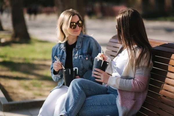 Zwei hübsche Frauen, die Fastfood essen. Schöne Mädchen auf Bank im Park sitzen und asiatisches Essen zu sich nehmen. Konzept Essen zum Mitnehmen — Stockfoto