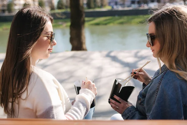 Zwei hübsche Frauen, die Fastfood essen. Schöne Mädchen auf Bank im Park sitzen und asiatisches Essen zu sich nehmen. Konzept Essen zum Mitnehmen — Stockfoto