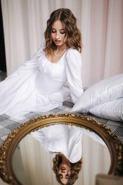 Κομψό γυναικείο μοντέλο κάθεται κοντά στον καθρέφτη με την αντανάκλασή της. Πανέμορφη και σέξι νύφη σε ένα κοντό φόρεμα στο κρεβάτι τεντώνει τα μαλλιά της — Φωτογραφία Αρχείου