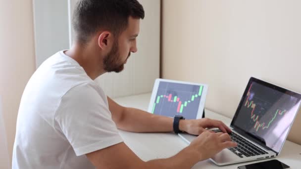 Hombre mirando gráfico de finanzas en el ordenador portátil y tableta, comerciante día, invertir desde casa. El joven supervisa sus inversiones. Hombre de negocios ambicioso — Vídeos de Stock