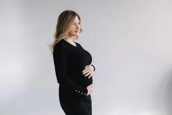 Élégante femme enceinte en robe noire tup mains sur le ventre et sourire. Fond blanc — Photo