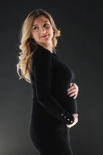 Средний отбор на беременную женщину кладет руки на живот. Женщина в черных платьях bodycon. Черный фон — стоковое фото