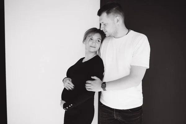 Donna incinta con il suo adorabile marito si abbracciano e mettono mano sulla pancia. Fotosessione in studio. Sfondo bianco e nero — Foto Stock