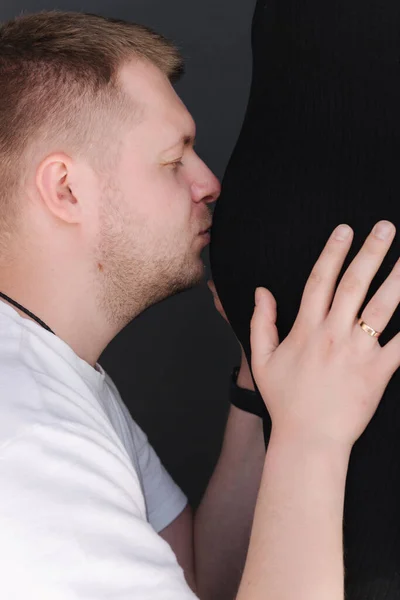 Un uomo gentile che bacia la pancia della moglie incinta. Uomo in camicia bianca e donna in abito nero. Da vicino. — Foto Stock