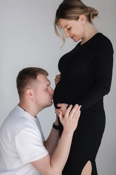 Jemný muž líbá svou těhotnou ženu na břiše. Muž v bílé košili a žena v černých šatech — Stock fotografie